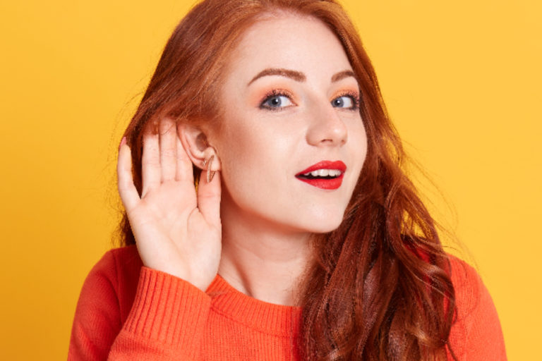 Funny astonished red haired European female, raising eyebrows, expressing surprise, keeping hand near ear trying to listen to gossips, wearing orange casual sweater.